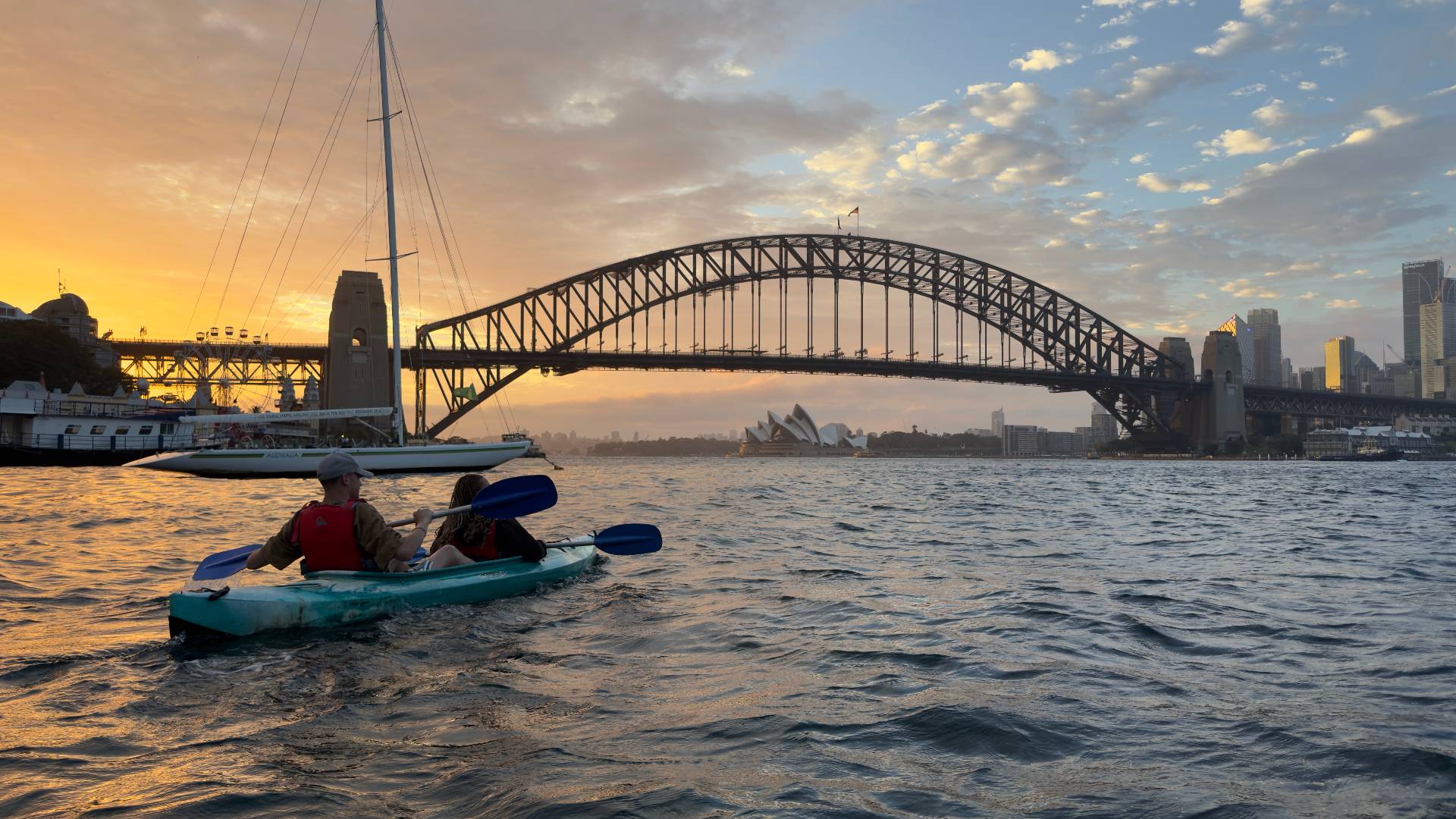 Couple paddling past sailboat with Sydney Harbour Bridge