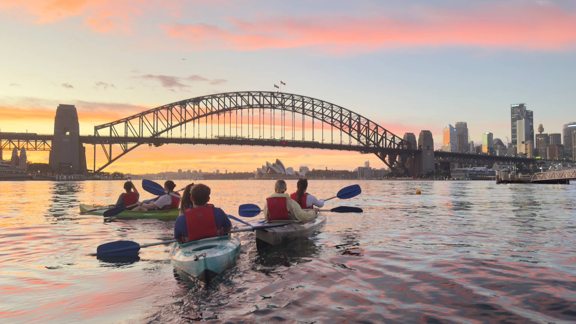 Group kayak tour paddling towards Sydney Harbour Bridge at sunset